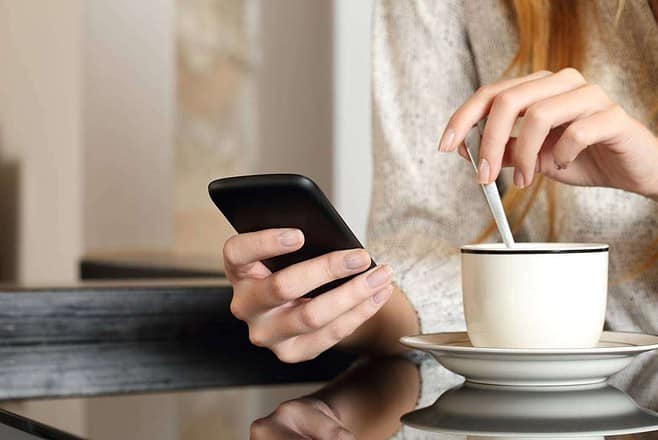 A woman using a cell phone while sitting at a table with a cup of coffee and reading an eBook on her tablette.