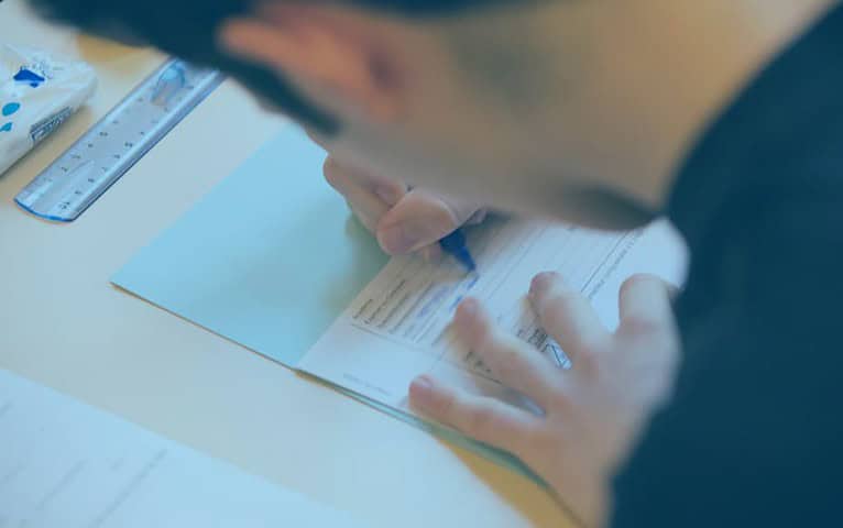 Une personne remplit un formulaire avec un stylo bleu sur un dossier ou un bloc notes bleu clair, travaillant peut être sur un essai en français. L'individu porte une chemise sombre. Une règle et une gomme sont visibles sur la table à côté du formulaire. L'accent est mis sur la main et la forme de la personne, le visage étant partiellement flou et flou.