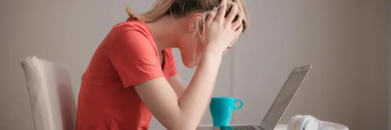 Une personne portant un t shirt orange est assise, penchée sur un ordinateur portable, les mains tenant sa tête en signe de frustration, probablement après avoir reçu des courriels de l'Éducation Nationale dont elle souhaite se désabonner. Une tasse turquoise et des écouteurs blancs sont posés sur la table à côté d'eux, sur un fond uni et clair.