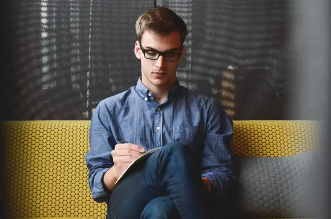 Un homme portant des lunettes et une chemise bleue est assis sur un canapé jaune à motifs, les jambes croisées, concentré sur la correction de ses notes dans un cahier. L'arrière plan présente un mur sombre et texturé.